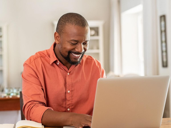 A man works at his laptop. He is smiling while wearing an orange shirt.
